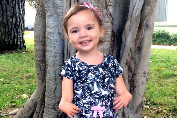 Smiling toddler girl with pink bow in her hair in front of tree