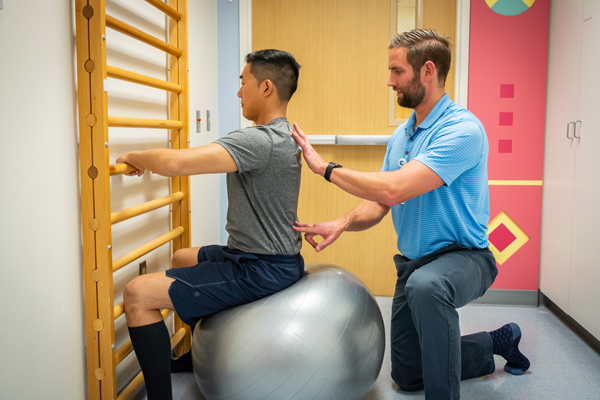 teen boy holding wall ladder, sitting on exercise ball, with male Schroth physical therapist