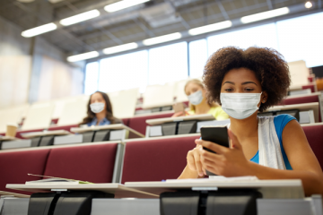 young woman in college class using phone wearing mask